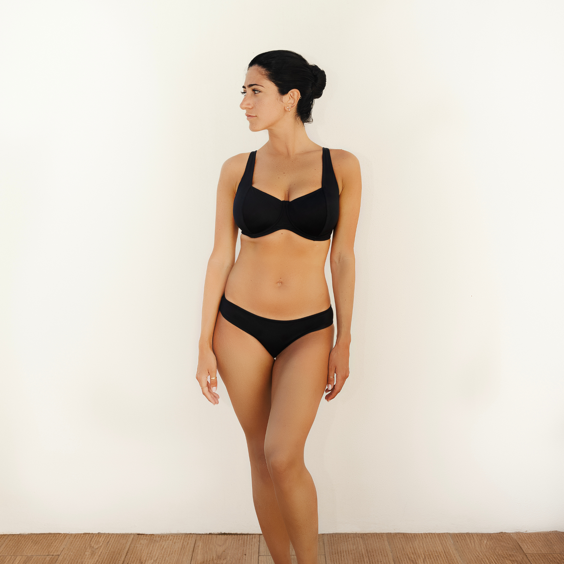 Black Balconette Shaped Swimwear snug bikini  top and classic bikini briefs worn by greek model looking at the side on a white backgrond